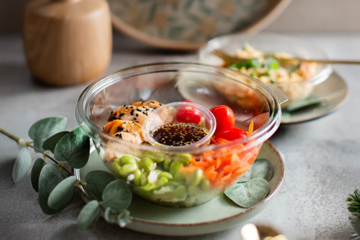 Kolorowa sałatka w plastikowej misce z łososiem, marchewką, pomidorkami koktajlowymi i sosem sojowym w małym pojemniczku, podana na talerzu w eleganckiej aranżacji.