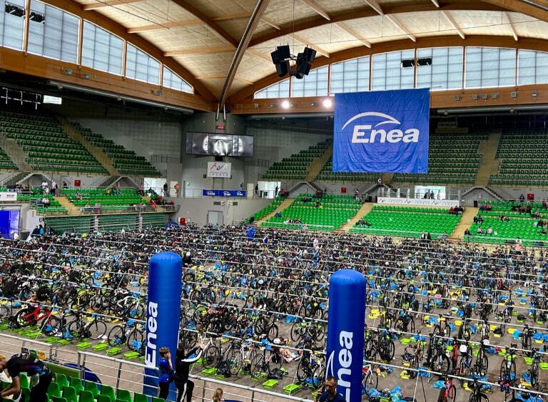 Hala sportowa z zielonymi trybunami i setkami rowerów w strefie zmian triathlonu, z dużym banerem Enea zawieszonym nad areną