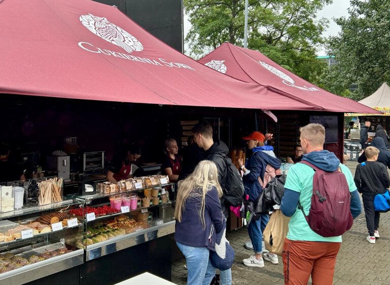 Stoiska Cukierni Sowa pod bordowymi namiotami z logo, z widoczną ladą pełną wypieków i klientów zamawiających desery oraz kawę